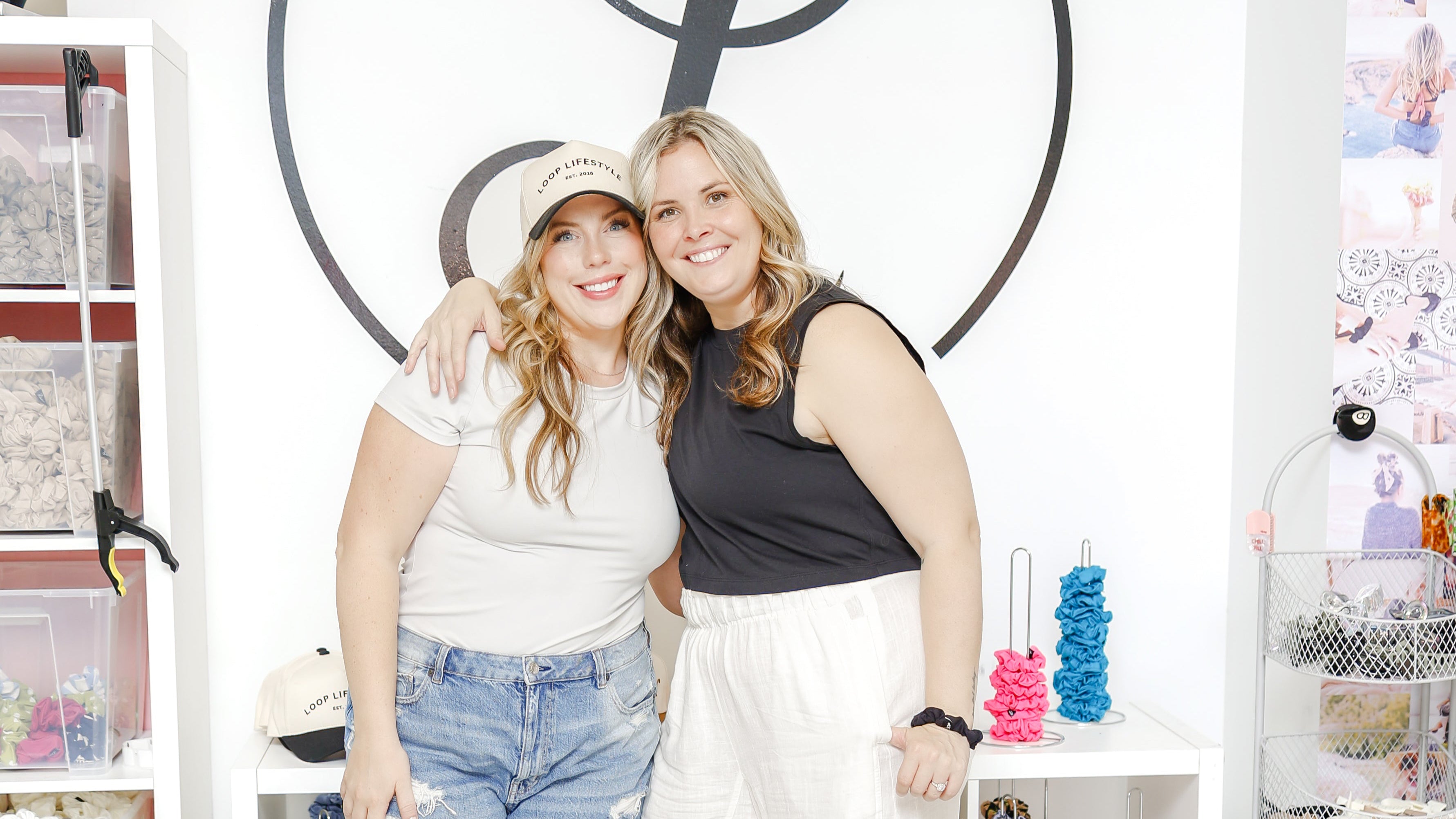 Two women posing in front of a 'Loop' branded wall with shelves displaying various items.